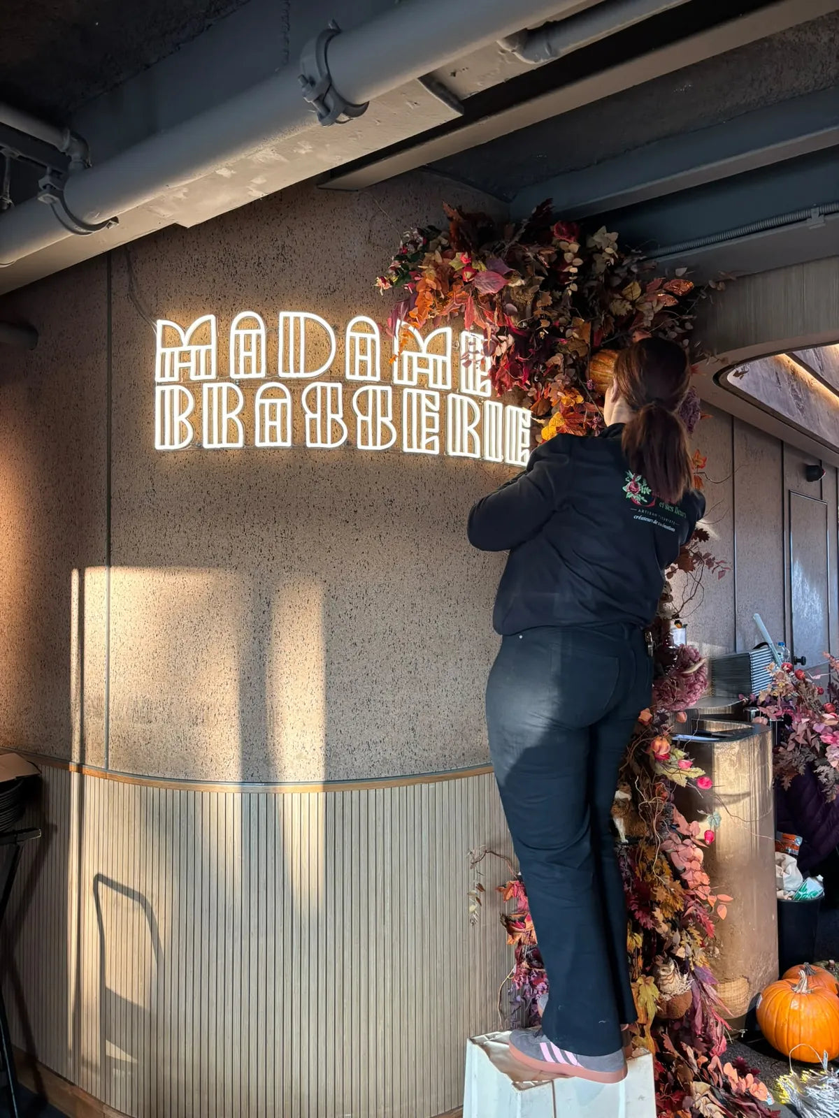 Une fleuriste de Camelia, sur un escabeau, accroche la guirlande de feuillages autour de l’enseigne Madame Brasserie