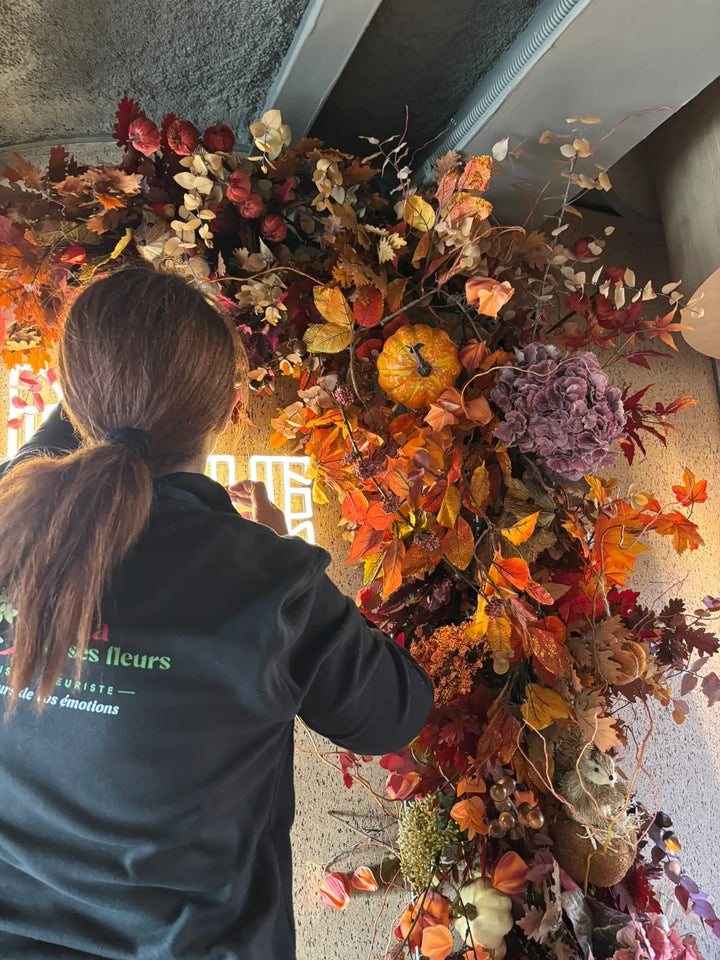 Une fleuriste de dos, logo Camelia et ses fleurs sur le sweat, pique des feuillages d’automne dans la guirlande