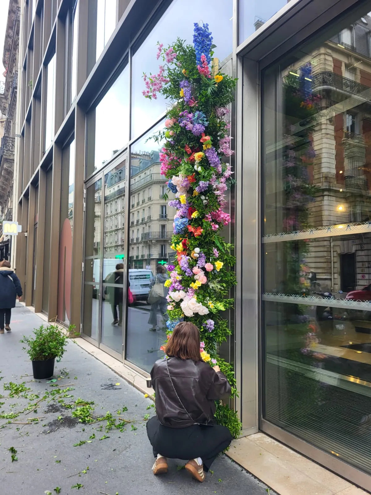 Une fleuriste accroupie monte une colonne de fleurs devant la façade vitrée