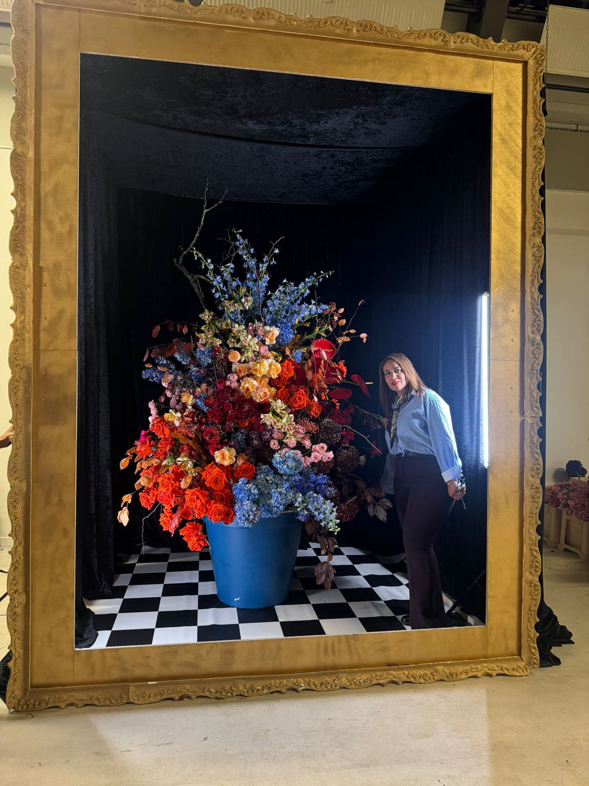 Kodsia Renaud à côté d’une grande composition florale présentée dans un cadre doré, sur un sol en damier